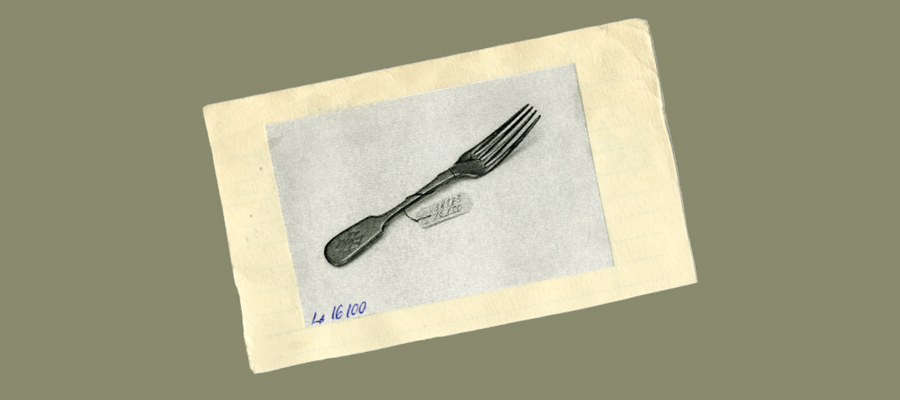 Black and white photograph of a metal fork with an intricate handle design mounted on off-white paper with the number 