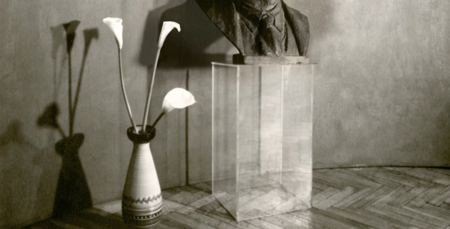 Sepia-toned photograph of a vase with three calla lilies on a wooden floor next to a transparent pedestal displaying a small sculpture.