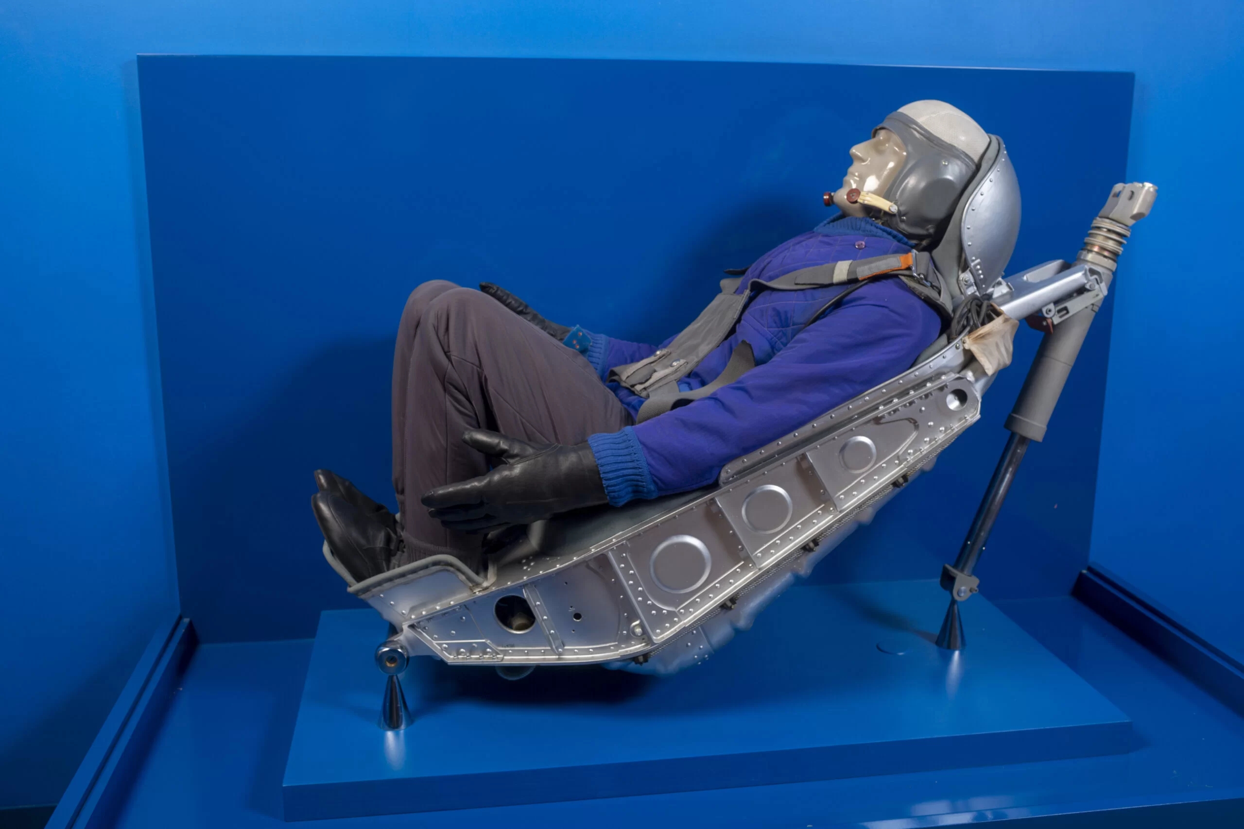 A spaceship's suspension chair with a male mannequin sitting in a blue jacket, dark pants, black shoes and black leather gloves.