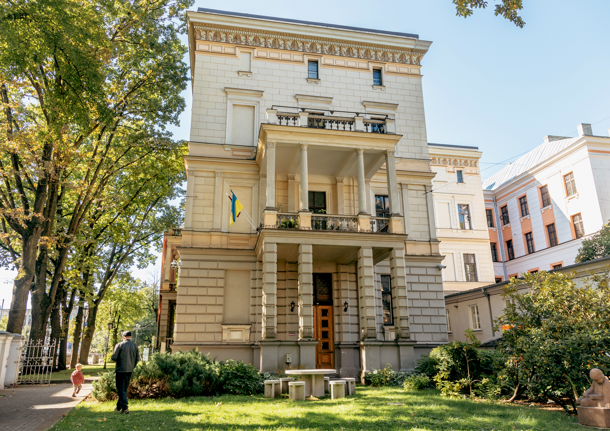 Image of the museum building. The museum has three floors, and the facade is in a light sand tone. In the foreground — the museum garden. On the lawn, there is a concrete table with chairs. On the left side of the museum, a green tree.