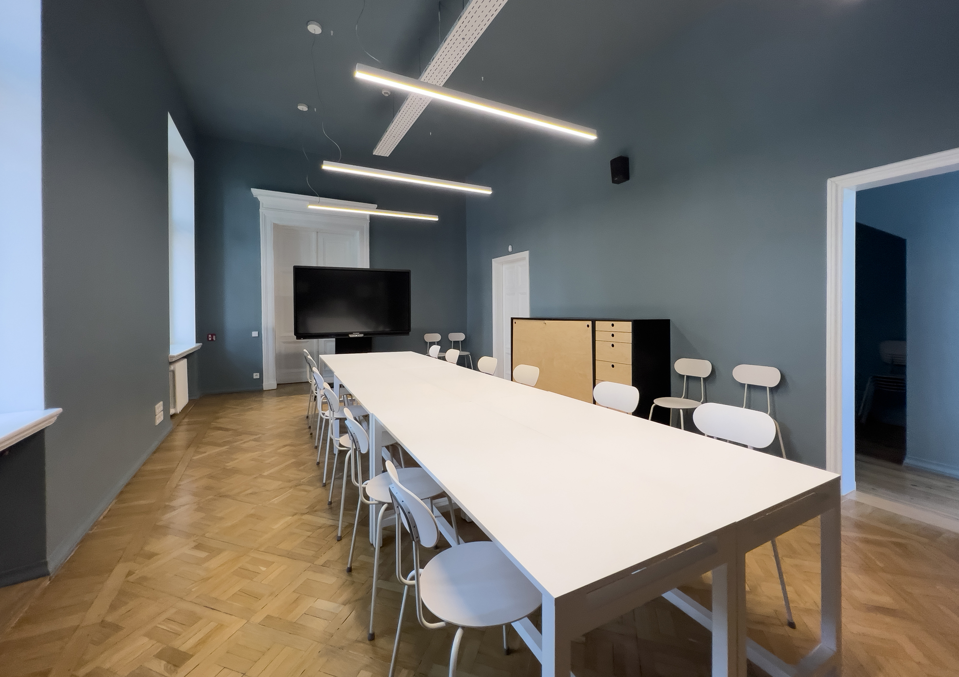Image of the third-floor meeting room. In the foreground, a rectangular white table surrounded by white chairs. At the far end of the room, a TV screen with white doors behind it. Light brown wooden floor, three ceiling lamps, and one bright window.
