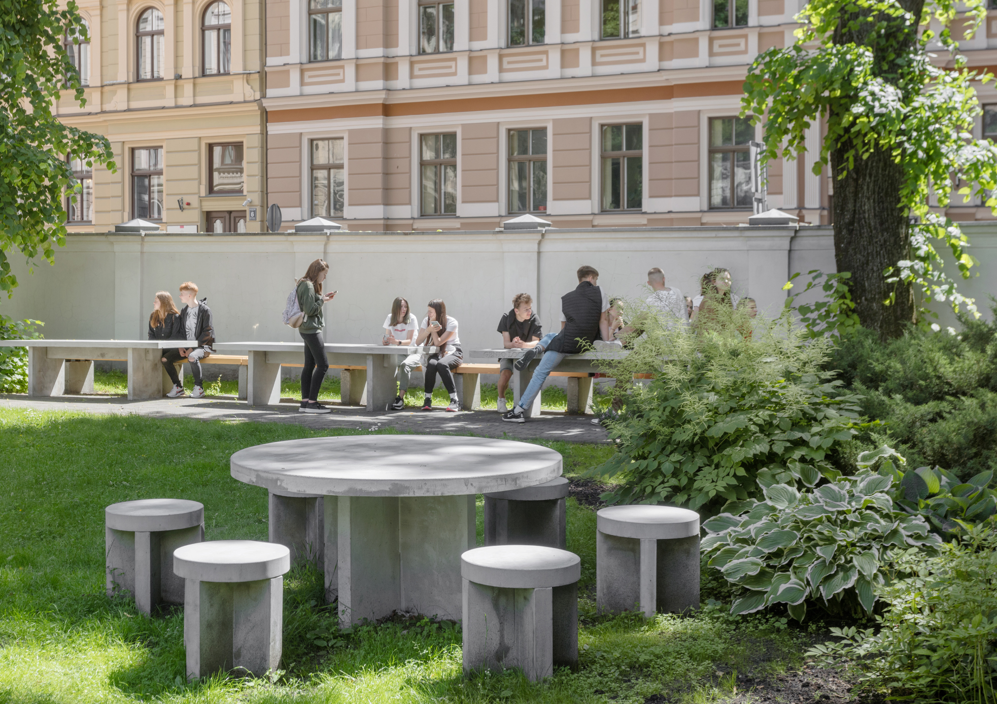 Museum garden. In the foreground is a circular concrete table with four chairs around it. In the background, green grass with trees and people sitting on concrete garden benches and tables.