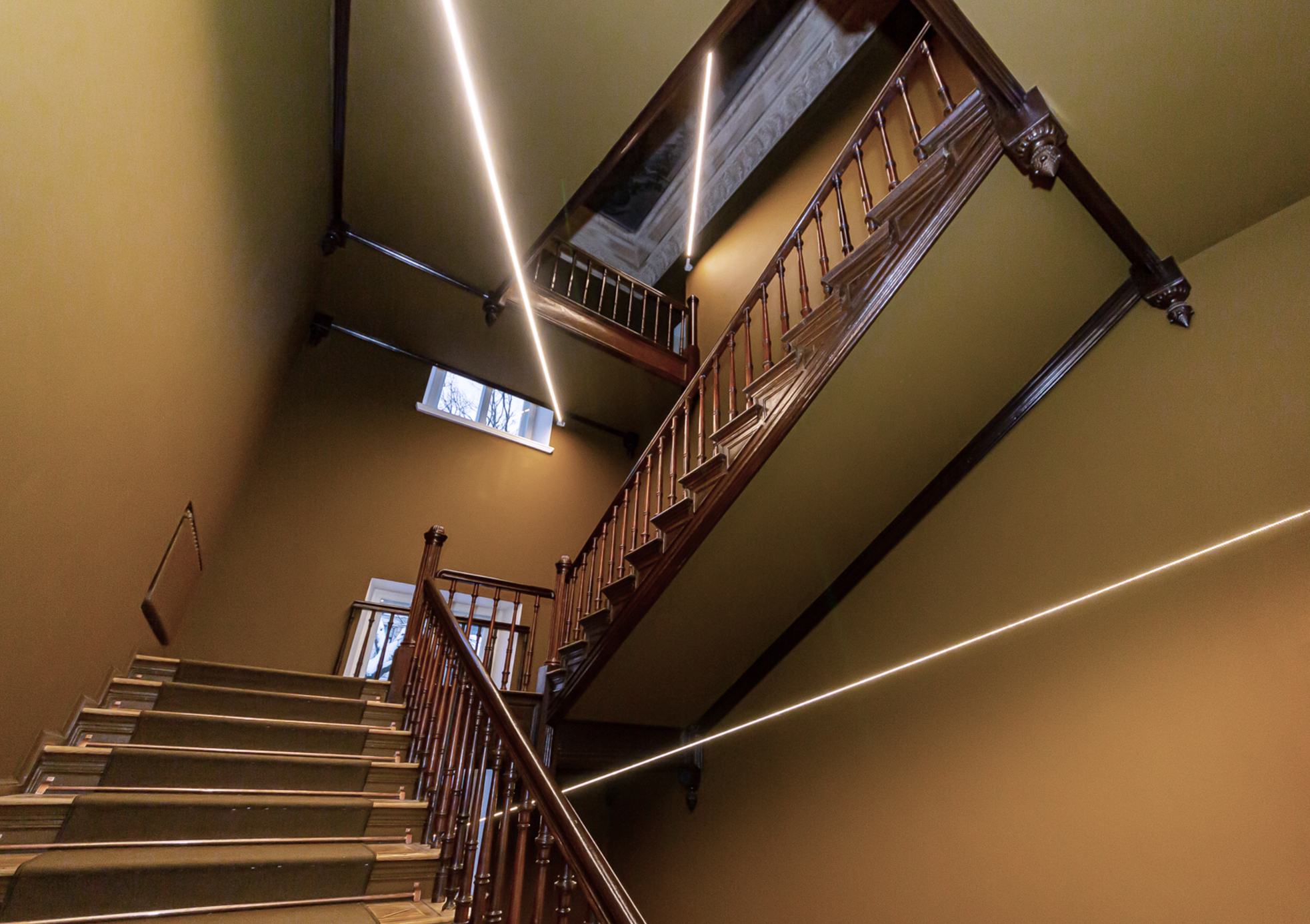 Image of the museum staircase. Brown walls and dark brown stair railings. Three lighting fixtures – LED cables.