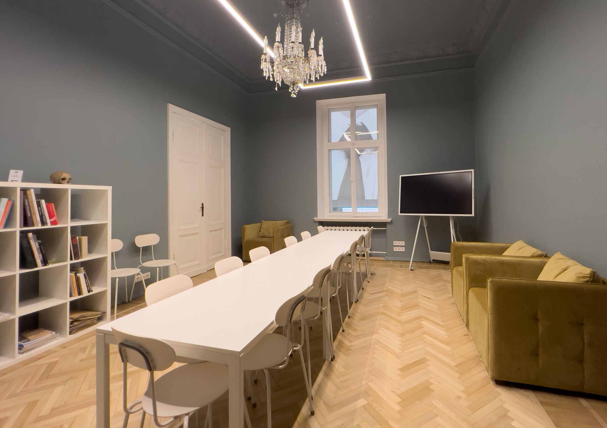 Image of the second-floor meeting room. Grey walls and ceiling, light wooden floor, a window. Door on the left. In the room interactive whiteboard and a white rectangular table with thirteen chairs around it. Against the right wall, there are two moss-green armchairs, and a white chandelier hangs from the ceiling.