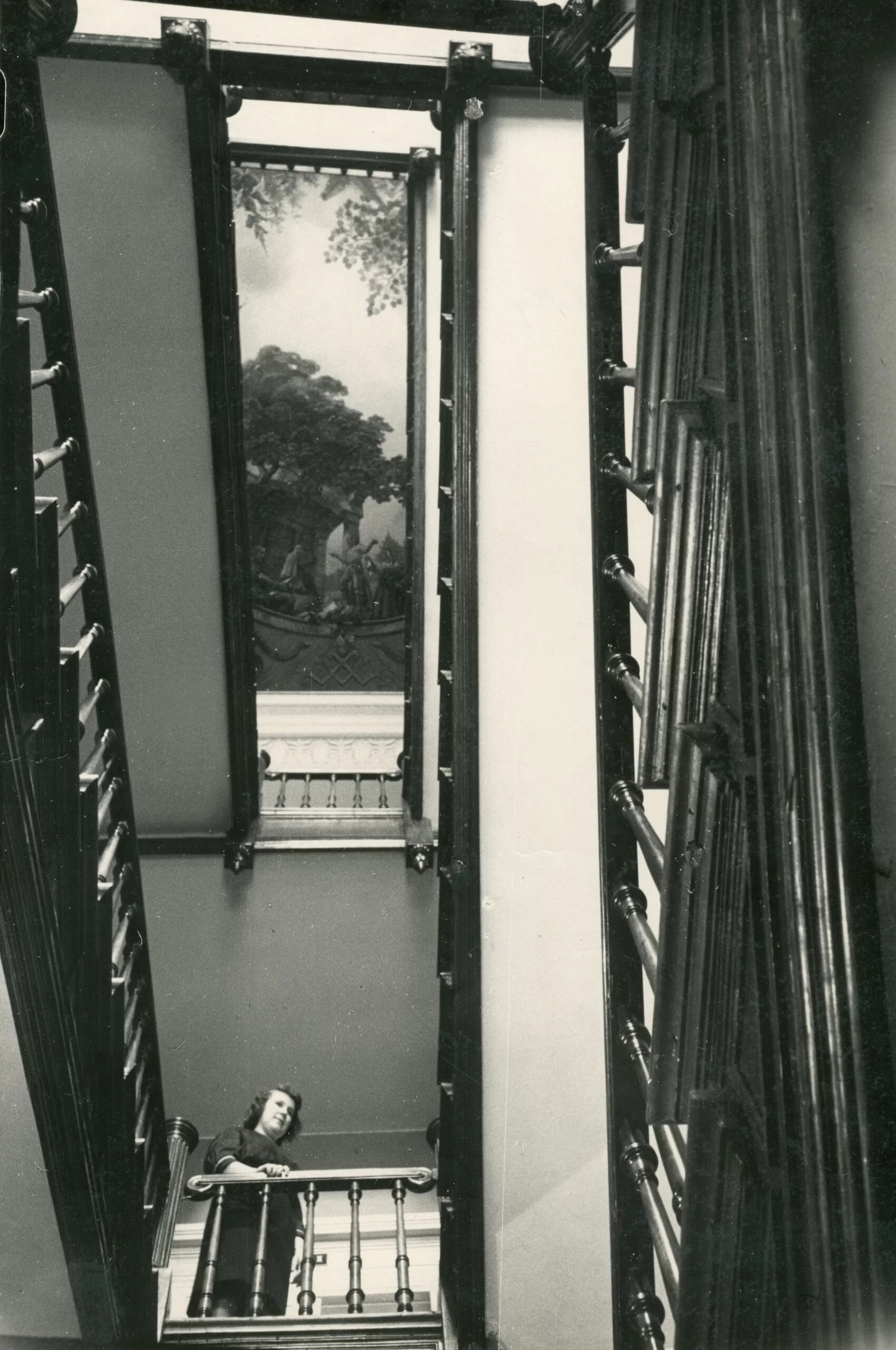 The museum's old stairs, photographed from the bottom looking up. At the very top you can see a ceiling painting with a view of nature. A woman can be seen on the stairs, holding on to a wooden railing.