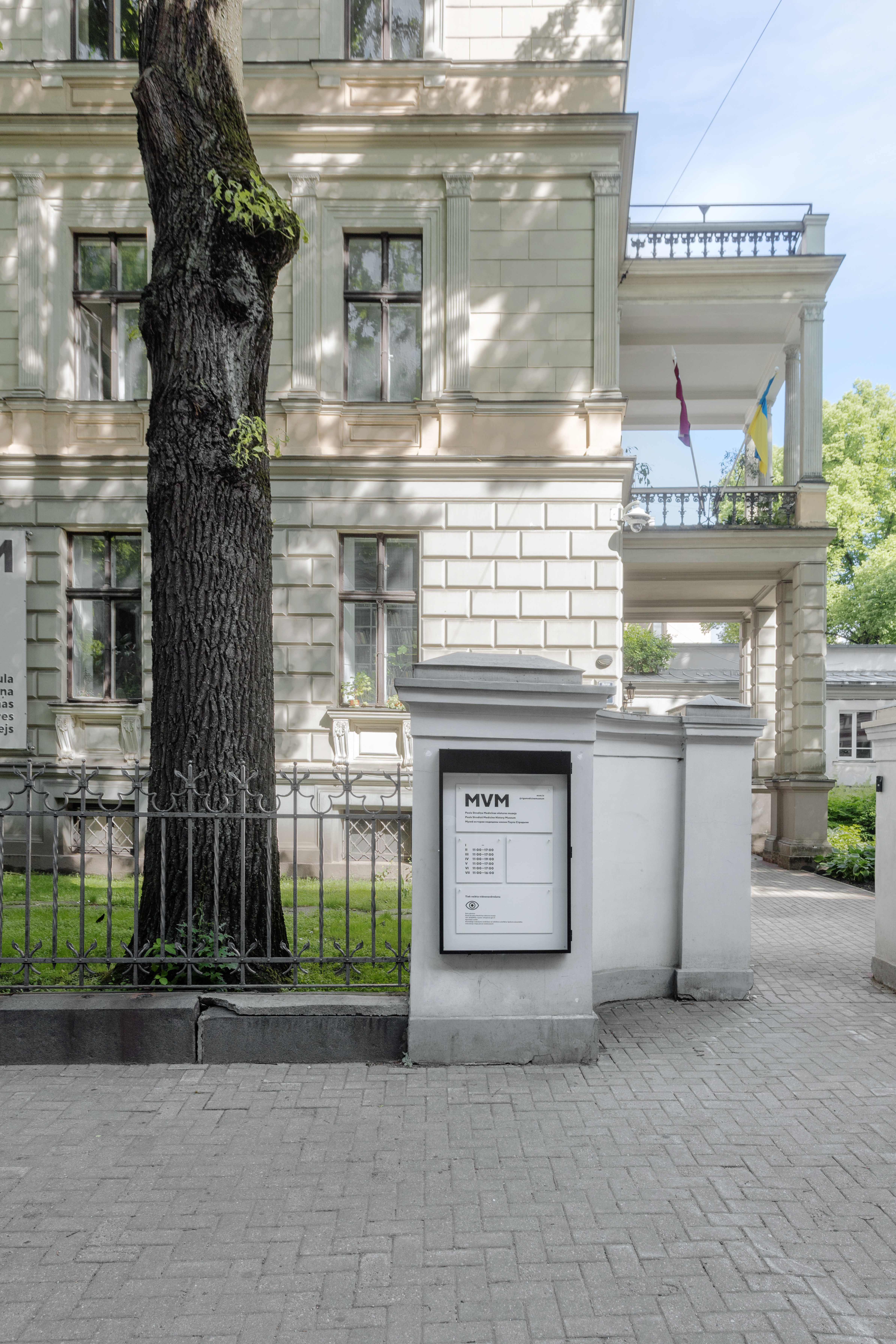 A street view of a beige building with white trim and a large tree in the foreground. There is an MVM sign board attached to the fence with a list of names and buzzers.