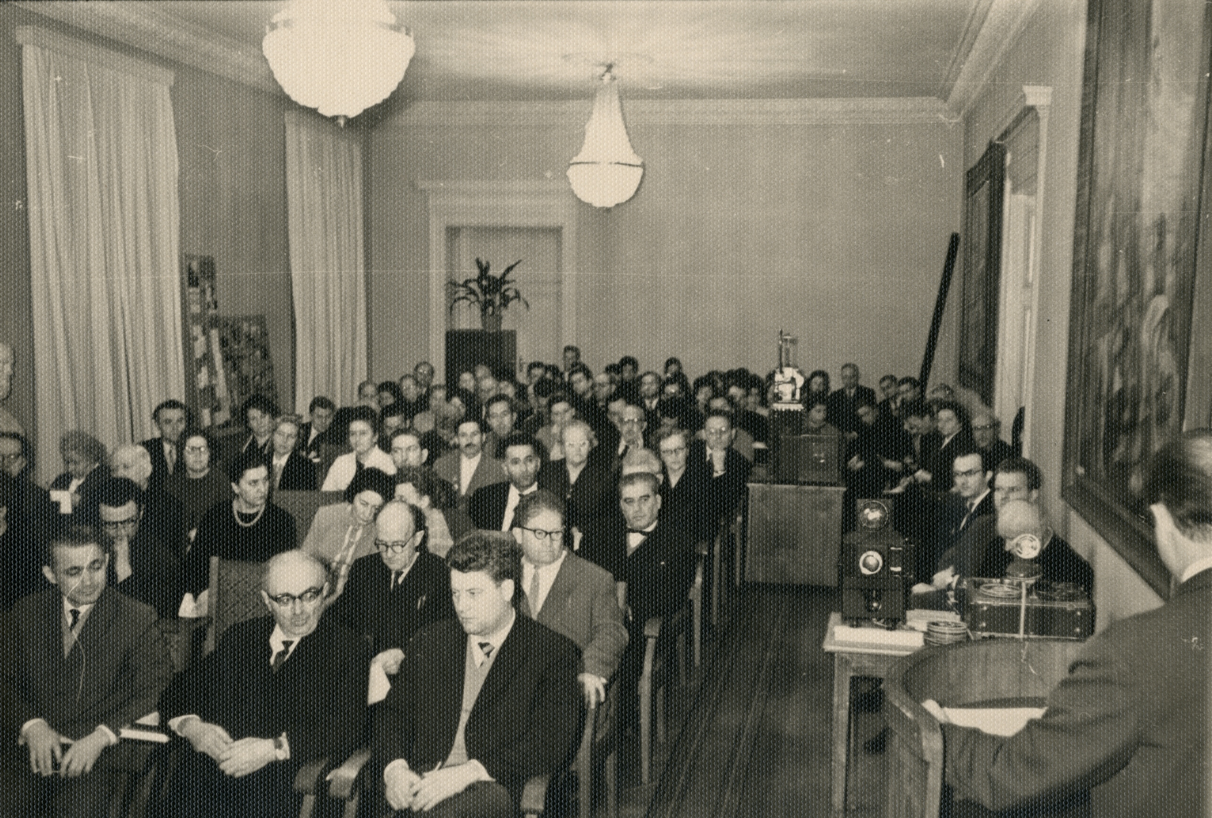 Black and white photo of a crowded room with many people sitting and facing forward, and a man standing next to a podium with a microphone and equipment, possibly giving a lecture or presentation. The room is decorated with framed pictures, long curtains and two hanging lamps.