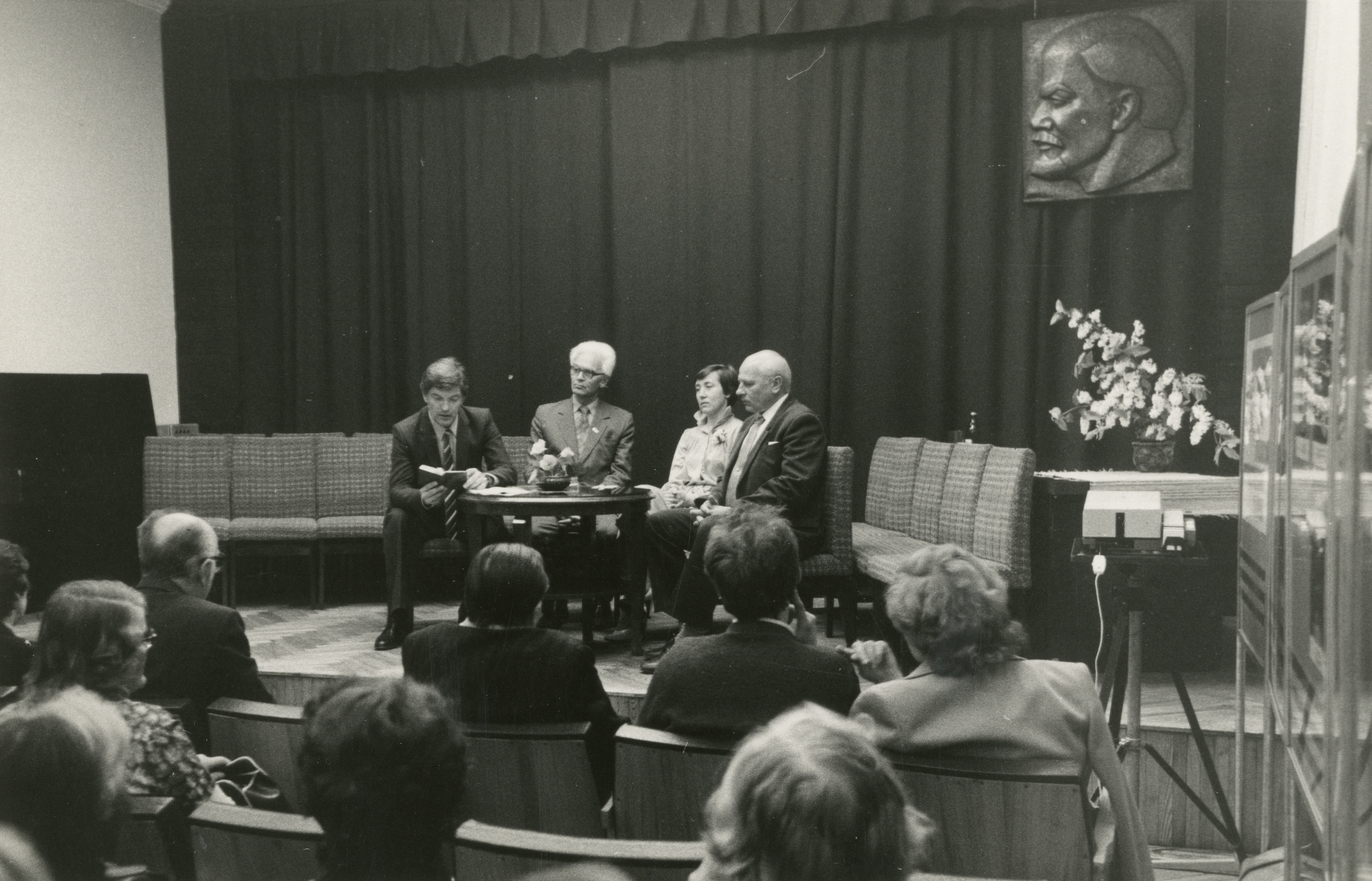 Black and white photo of four people sitting on a stage with a small table in front, in discussion mode, facing the audience in a room with curtains as a background and paintings on the wall. The audience seems to be listening intently. On the right is a display case a