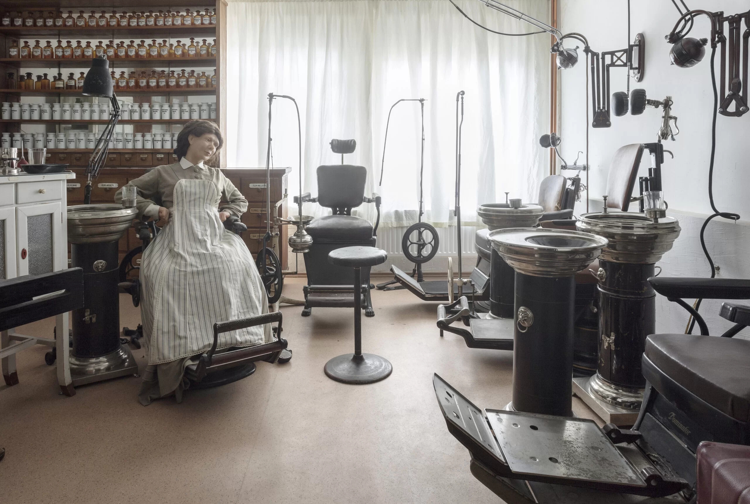 A photograph from a museum exhibit showing a female mannequin sitting on a medical chair. Around her are various medical chairs and other devices. As well as a medical shelf.