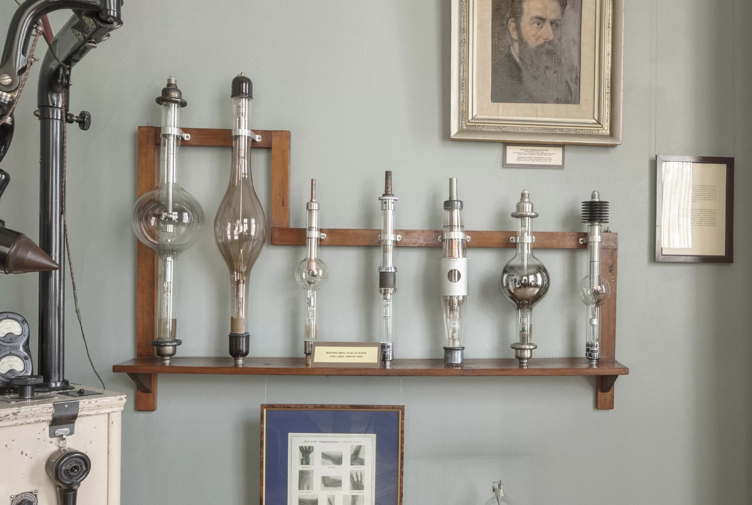 Various X-ray lamps from the 20th century are placed on the wall. A portrait of a man and other informative tablets can also be seen nearby.