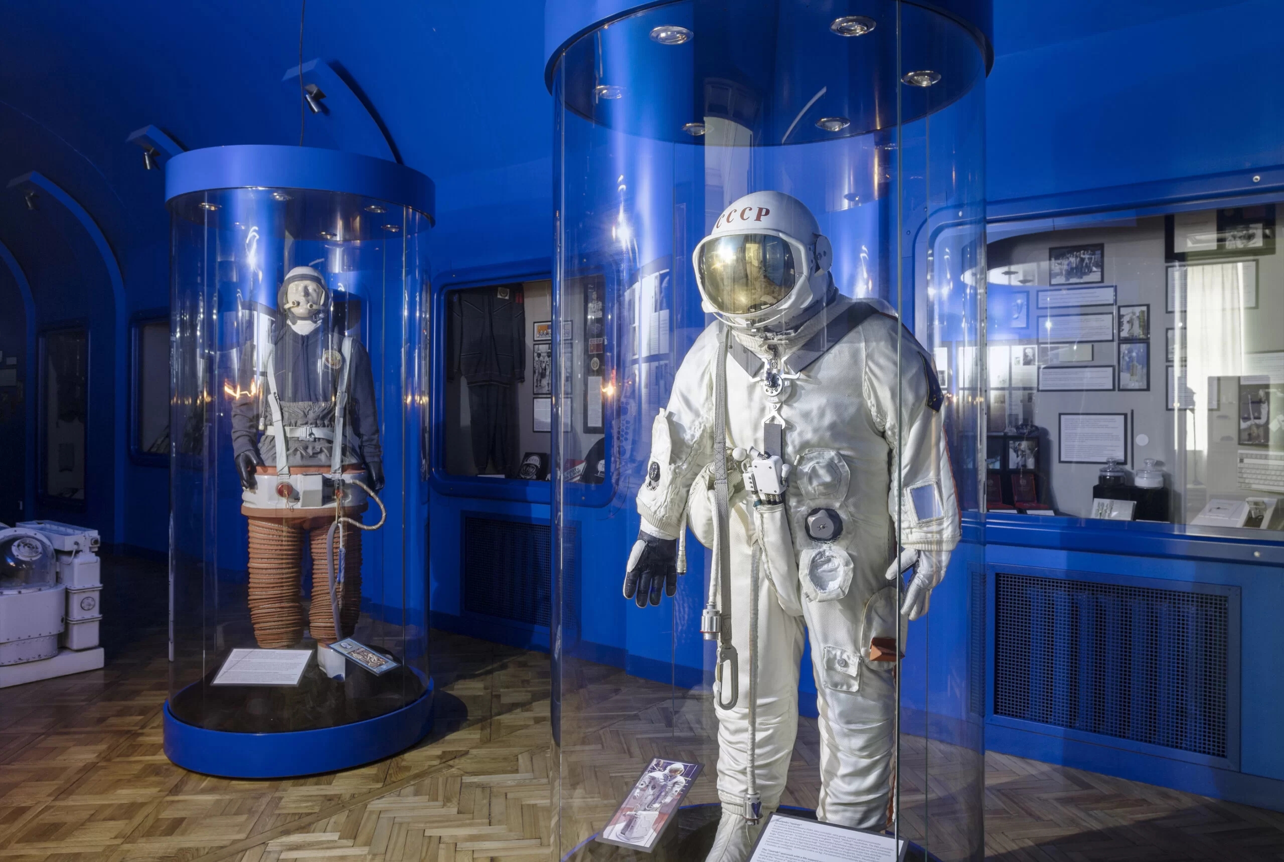A photo from the museum's space exhibit showing two mannequins dressed as astronauts. In the background are blue walls on which there are various informative tablets and exposition windows with exhibits.