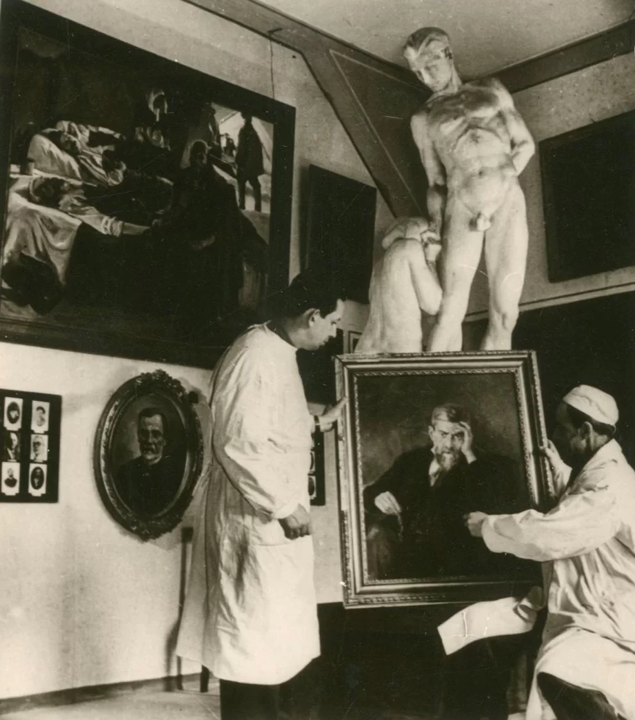 A photograph from the installation of the exposition showing two people holding a portrait. In the background is a large white human sculpture and other paintings on the walls.