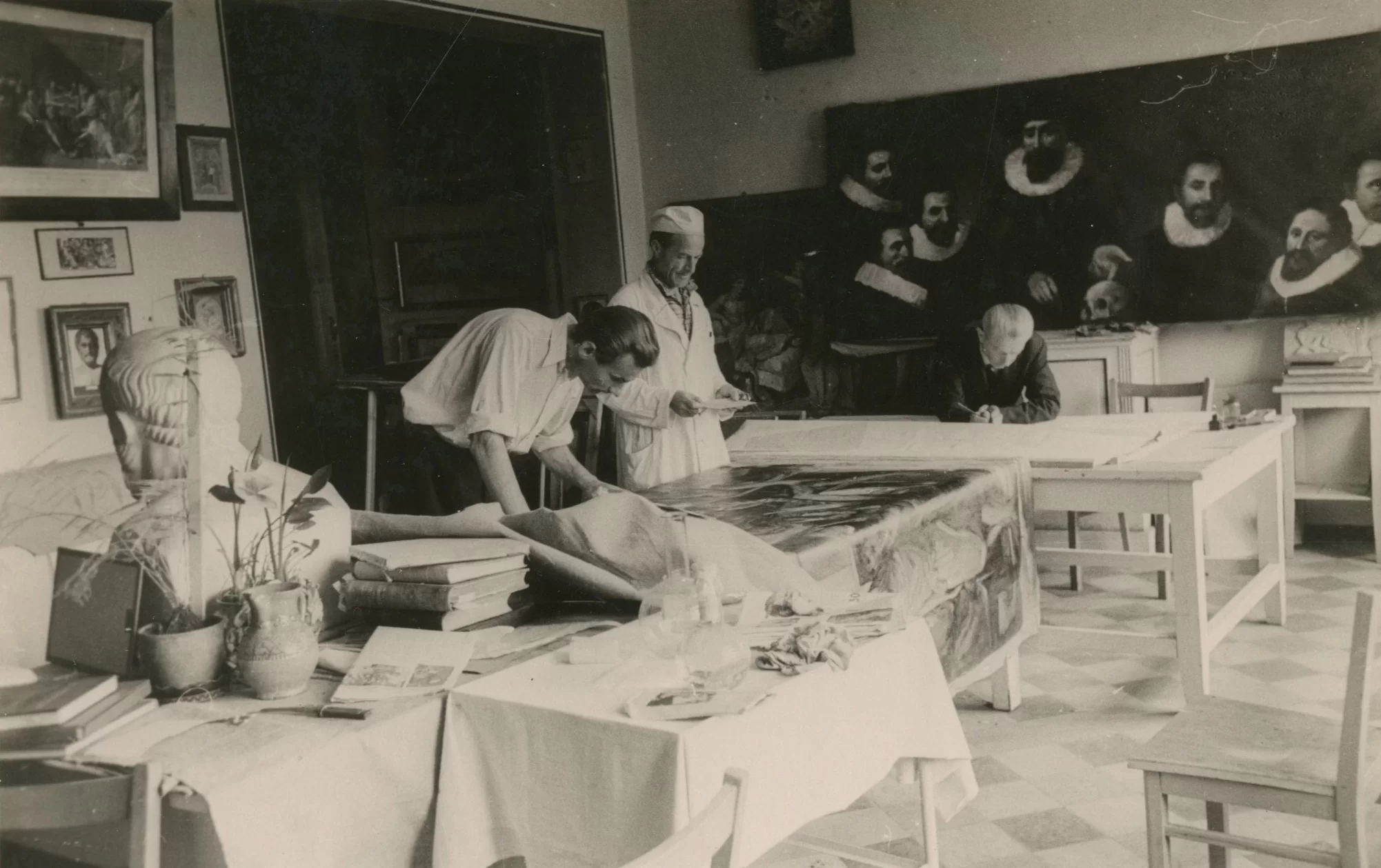 A photograph from the installation of the exposition showing people studying a painting at tables. Other paintings on the walls can be seen in the background.