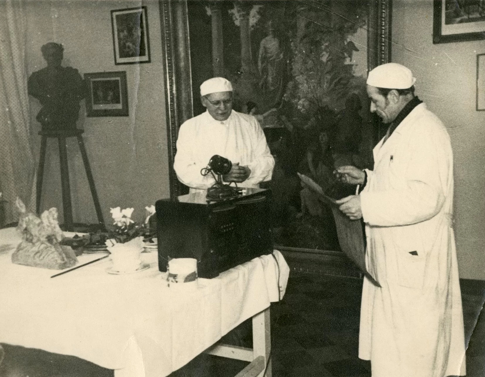 A photo of the exposition installation showing two people next to a voice recording device. There are some exhibits on the table and several paintings on the walls.
