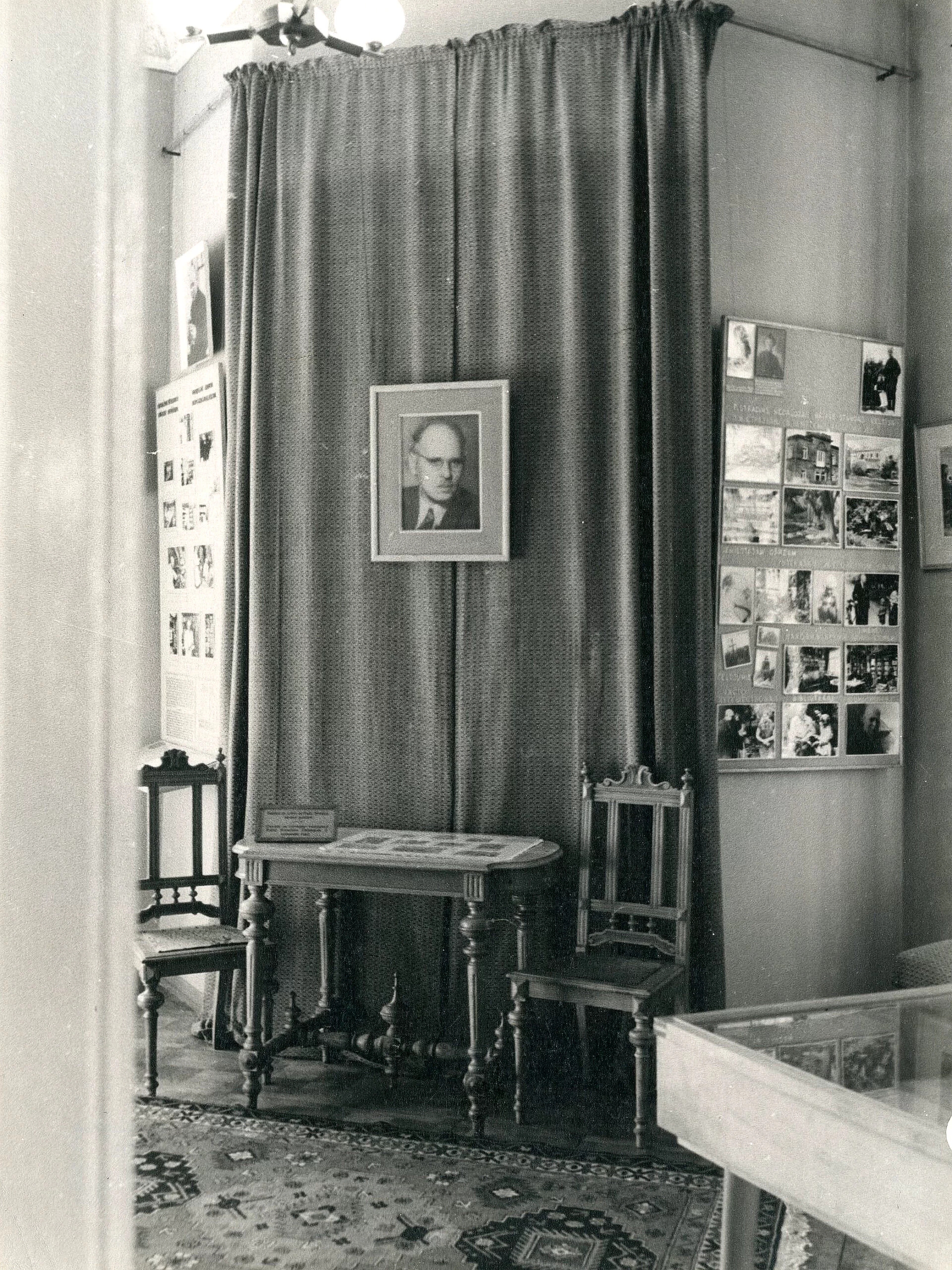 Photo of one of the museum exhibition rooms. Several photos can be seen around, in the middle there is a table with two chairs, above which is a photo of Pauls Stradiņš.
